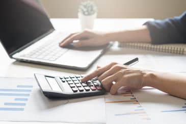 A person's left hand is operating a calculator while their right hand is on a laptop keyboard.