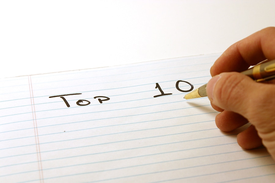 A person's hand writing "TOP 10" on the top line of a lined notebook page