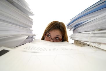 A young woman with glasses is looking up from behind stacks of paper.
