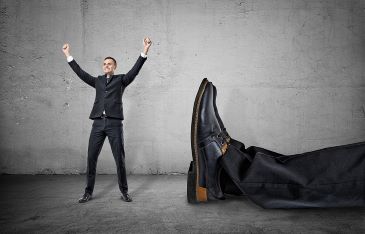 A small businessman standing with his arms up and smiling happily near a giant leg in trousers and shoe lying near him, on the concrete gray background.