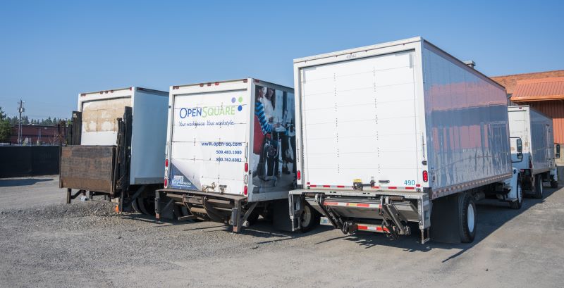 Large delivery trucks with rear liftgates are parked side-by-side in the yard of an industrial facility.