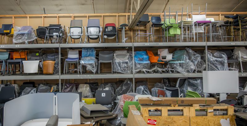 Steel shelving in a warehouse is stacked with many different styles and colors of office chairs.