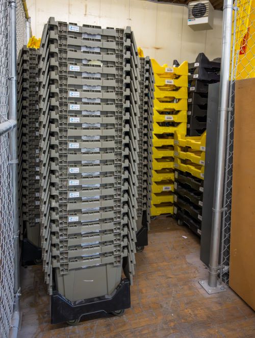 Wheeled dollies stacked with lidded plastic crates are waiting in a fenced section of a warehouse.
