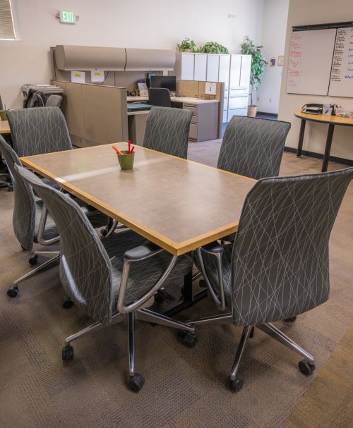 Six rolling office chairs sit around a conference table in an open area of an office with desks and file cabinets in the background.