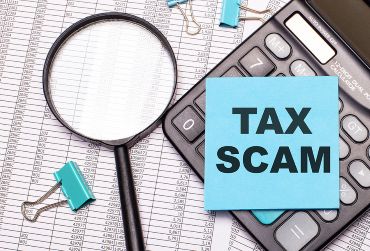 On the table are reports, a magnifying glass, a calculator, and a blue note sticker with the word TAX SCAM.