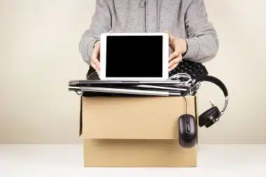Person standing behind a cardboard box full of old computers, phones, and devices that are ready for recycling.