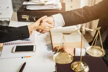 Two legal professionals are shaking hands across a desk cluttered with legal documents.