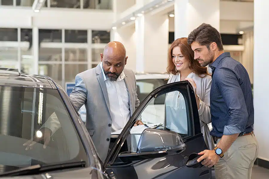 A car salesman is showing all the car features to a young couple in a showroom.