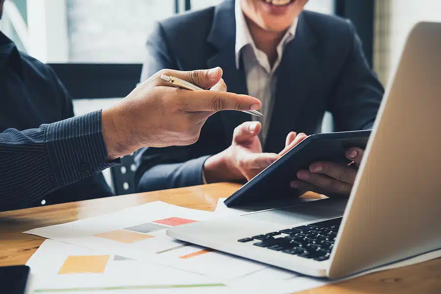 Colleagues in business attire discussing printed charts, a tablet, and a laptop on a wooden table
