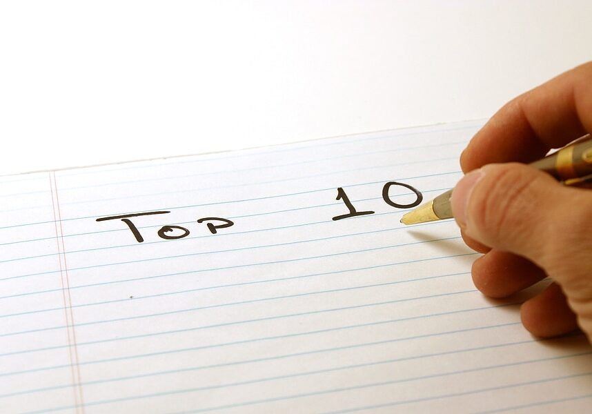 A person's hand writing "TOP 10" on the top line of a lined notebook page