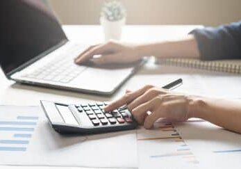 A person's left hand is operating a calculator while their right hand is on a laptop keyboard.