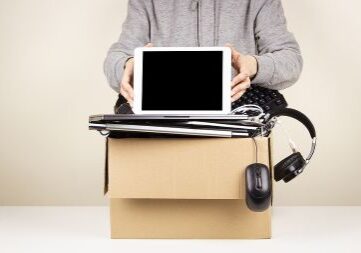 Person standing behind a cardboard box full of old computers, phones, and devices that are ready for recycling.