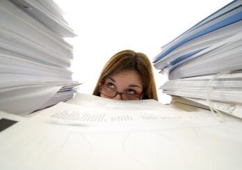 A young woman with glasses is looking up from behind stacks of paper.